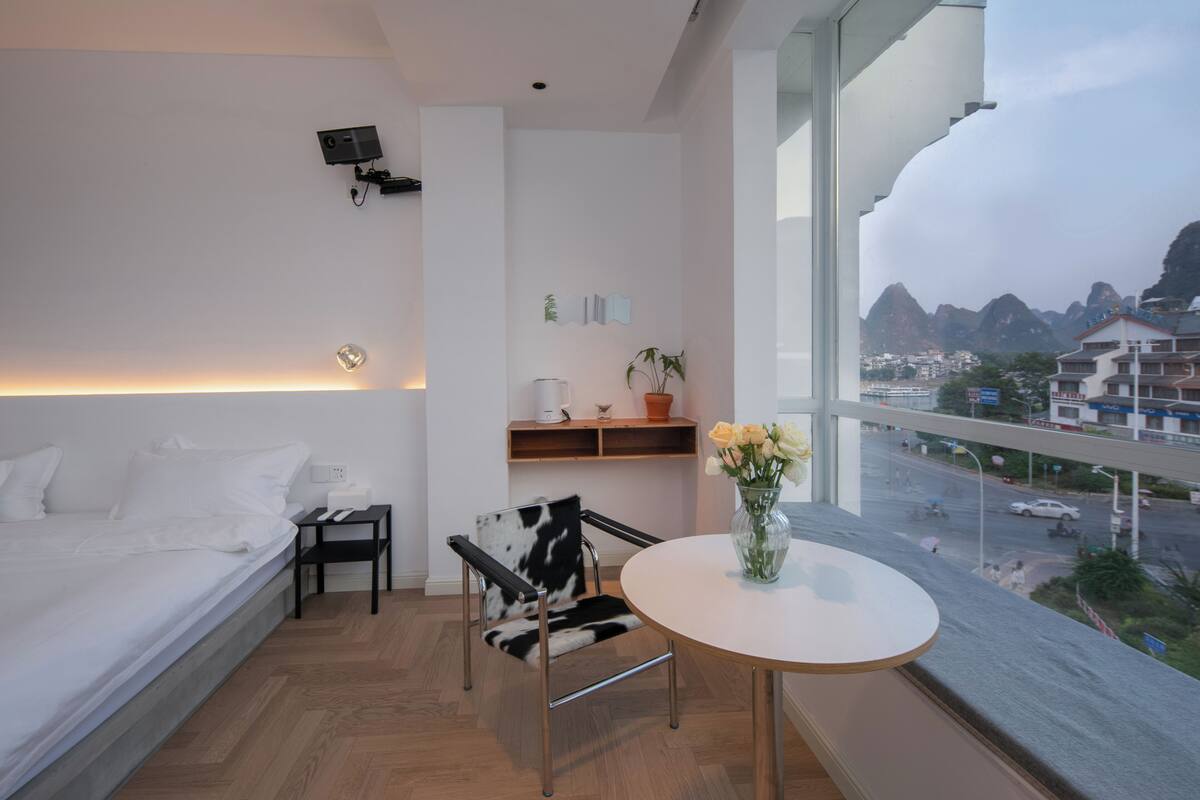 A modern hotel room with a bed, a small table with a vase of flowers, a chair, and a view of a cityscape with mountains.