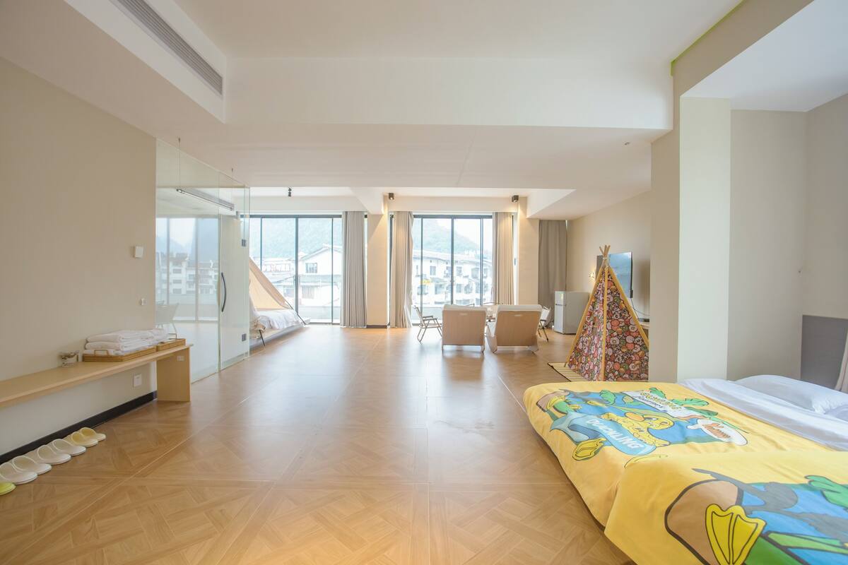 A hotel room with a bed, a desk, a chair, and a large window offering a view of buildings outside.