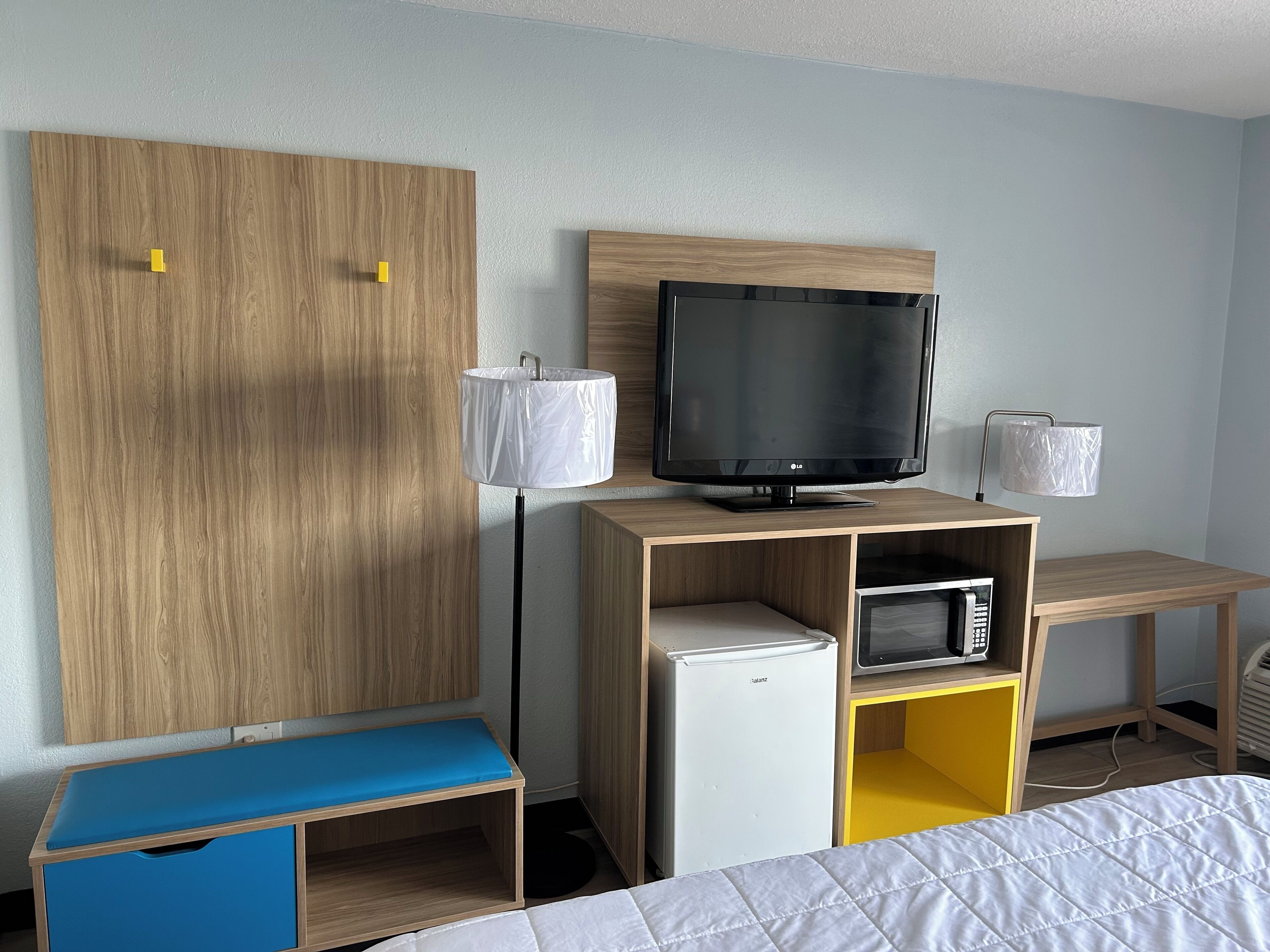 A hotel room with a wooden wardrobe, a television on a wooden stand, a small refrigerator, a blue bench, and a bed with a white quilt.