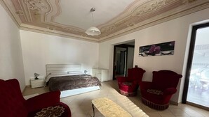 A bedroom with a bed, two red armchairs, a small table, a nightstand, and a painting on the wall. - Lercarappart 1 (Lercara Friddi)