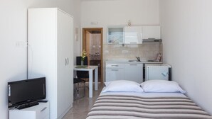 Ein kleines Hotelzimmer mit Bett, Fernseher, Schreibtisch und einer Küchenzeile.