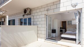 A modern apartment with a balcony, a sliding glass door leading to a bedroom with a bed and a desk, and a kitchenette area.