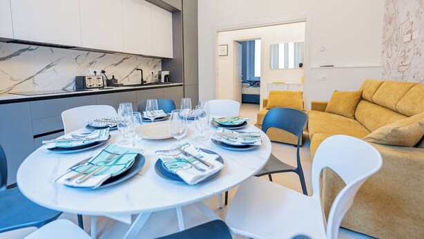 Uma sala de estar moderna com área de jantar preparada para quatro pessoas, apresentando uma mesa redonda, cadeiras brancas e um sofá amarelo.