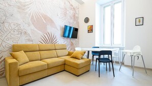 Uma sala de estar moderna com um sofá amarelo, uma televisão de tela plana, um espaço de refeições com mesa e cadeiras, e uma janela grande.