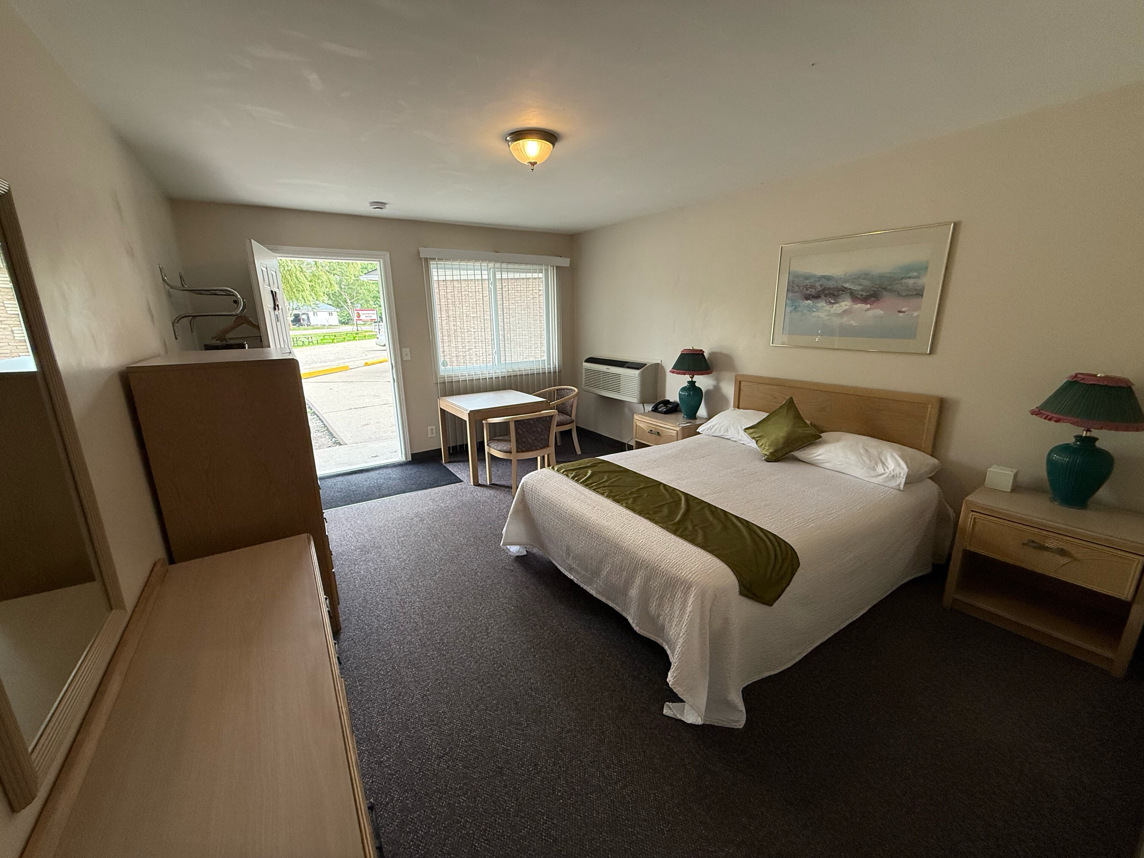 Een hotelkamer met een bed, nachtkastje, lamp, stoel, bureau en een schilderij aan de muur.