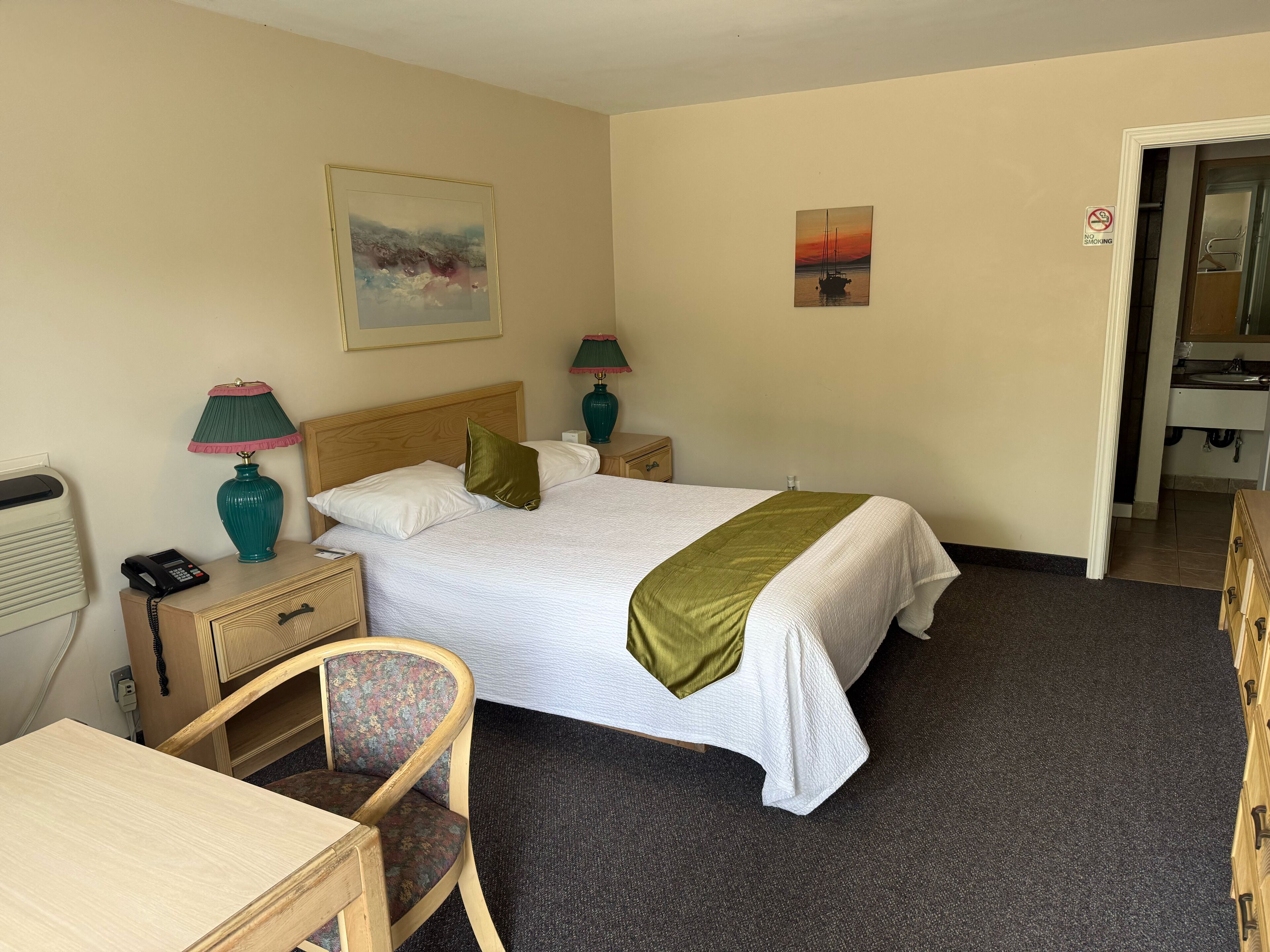 A hotel room with a bed, two bedside tables with lamps, a chair, a desk, a painting on the wall, and a bathroom visible through an open door.
