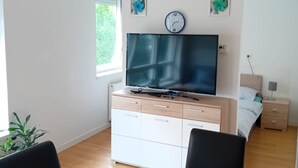Un dormitorio moderno con una televisión de pantalla plana, una mesita de noche con lámpara, una ventana con persianas y una planta en maceta.