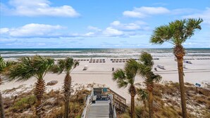 Vista de una playa con una escalera de madera que baja a la arena, palmeras y sillas de playa.