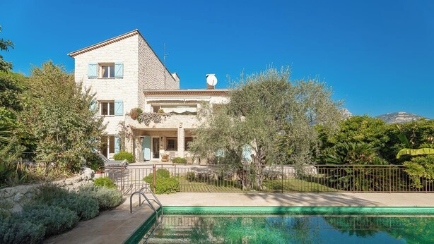 A two-story stone house with a swimming pool, surrounded by lush greenery and a clear blue sky.