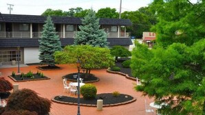 A courtyard with a brick pathway, surrounded by various trees and shrubs, and a building with a balcony in the background. - Avalon Inn (Sandusky)