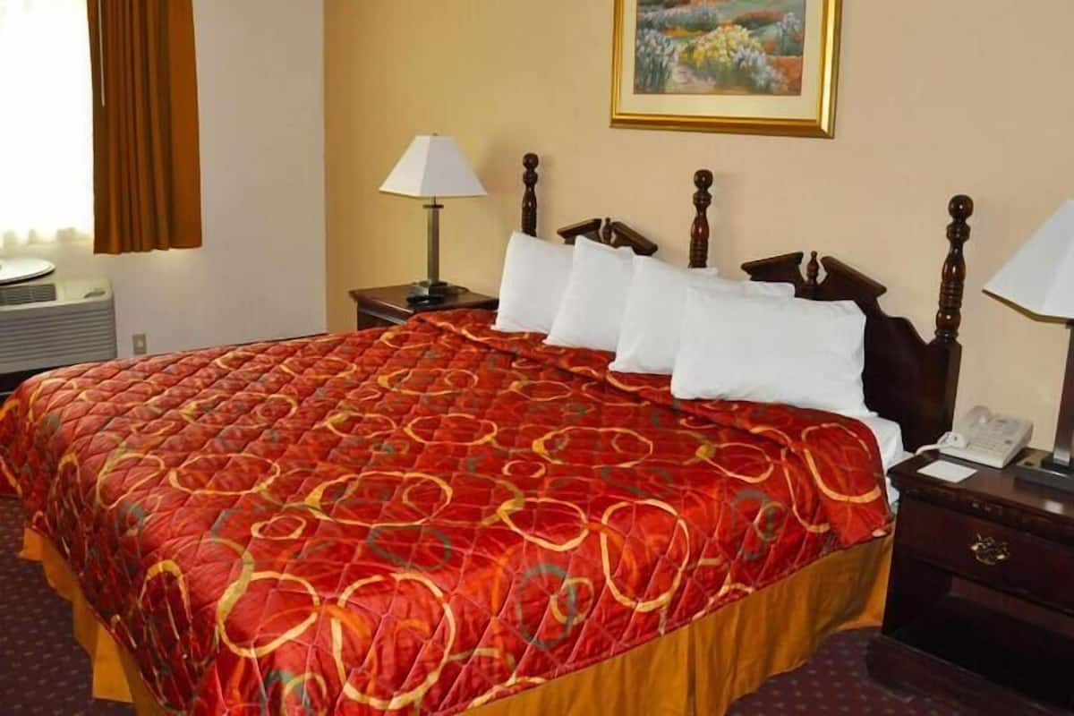Quarto de hotel com uma cama grande, uma mesinha de cabeceira com abajur, um quadro na parede e uma janela com cortinas.