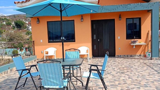 An outdoor patio with a table and chairs, a blue umbrella, and a house with orange walls and a tiled roof.