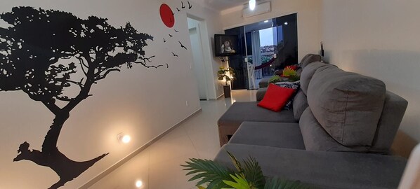 A modern living room with a grey sofa, a red cushion, a wall decal of a tree, and a TV on a stand.