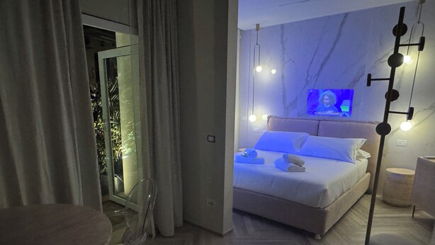 Habitació d'hotel moderna amb un llit, taules de nit, un mirall i vista a un balcó amb plantes.