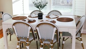 A dining area with a white table and wicker chairs, a chandelier, and a view through large windows. - Charming Scarborough Beach, Narragansett 3-bedroom home getaway (Narragansett)