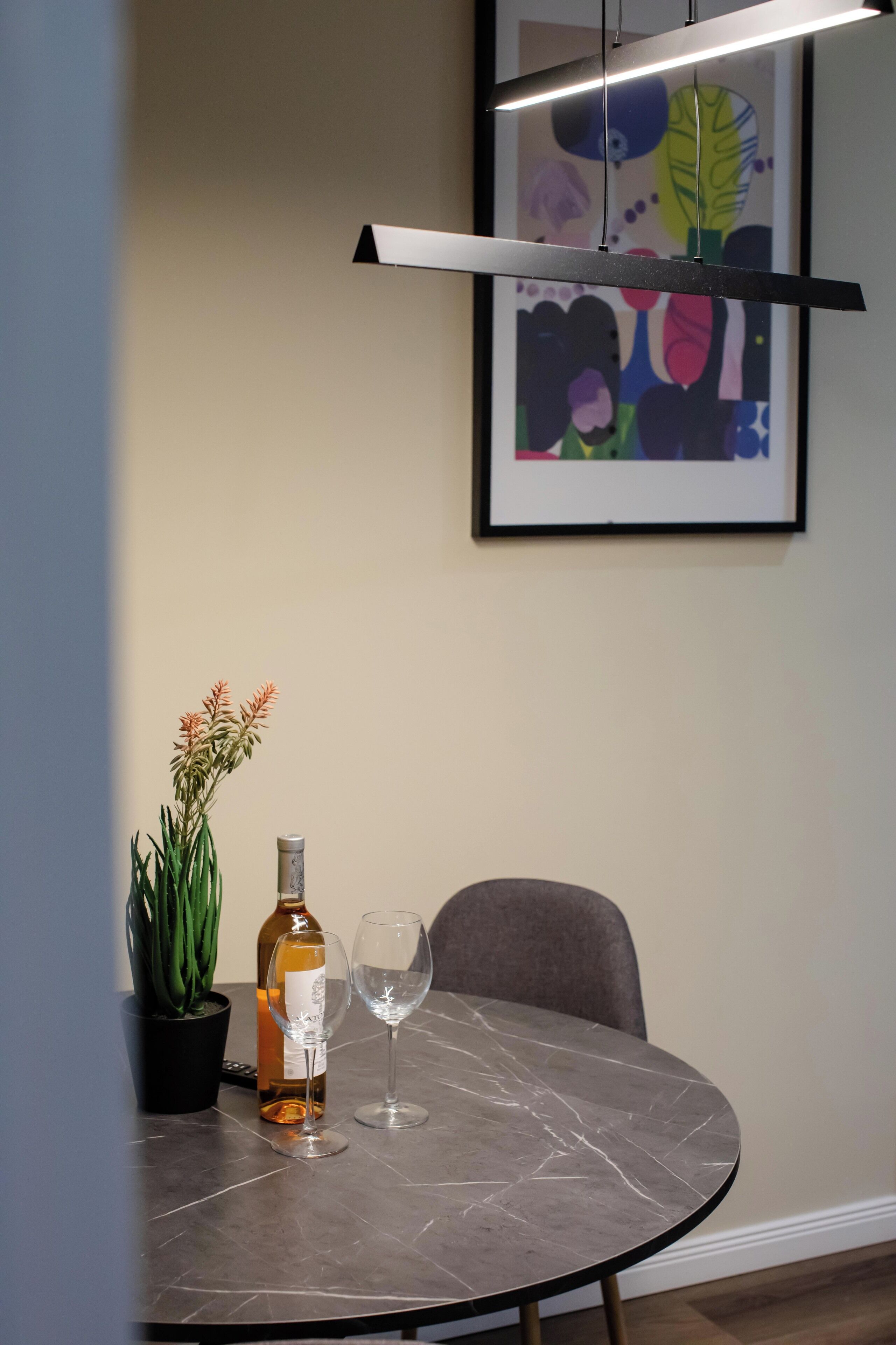 Una mesa de comedor con una botella, una copa de vino, una planta en maceta y una obra de arte enmarcada en la pared.