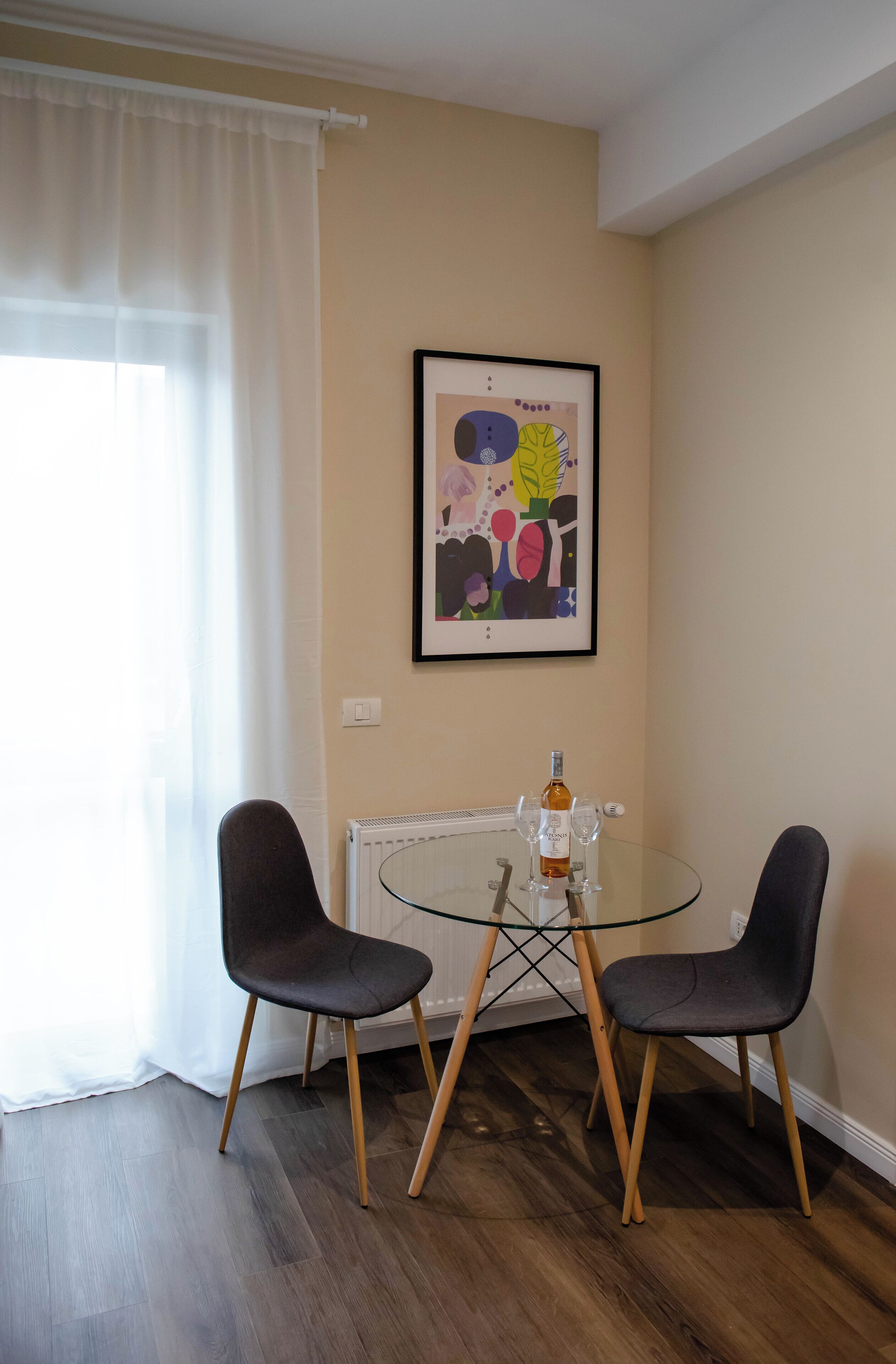 Zona pranzo con tavolo in vetro, due sedie, un termosifone e un quadro astratto incorniciato al muro.
