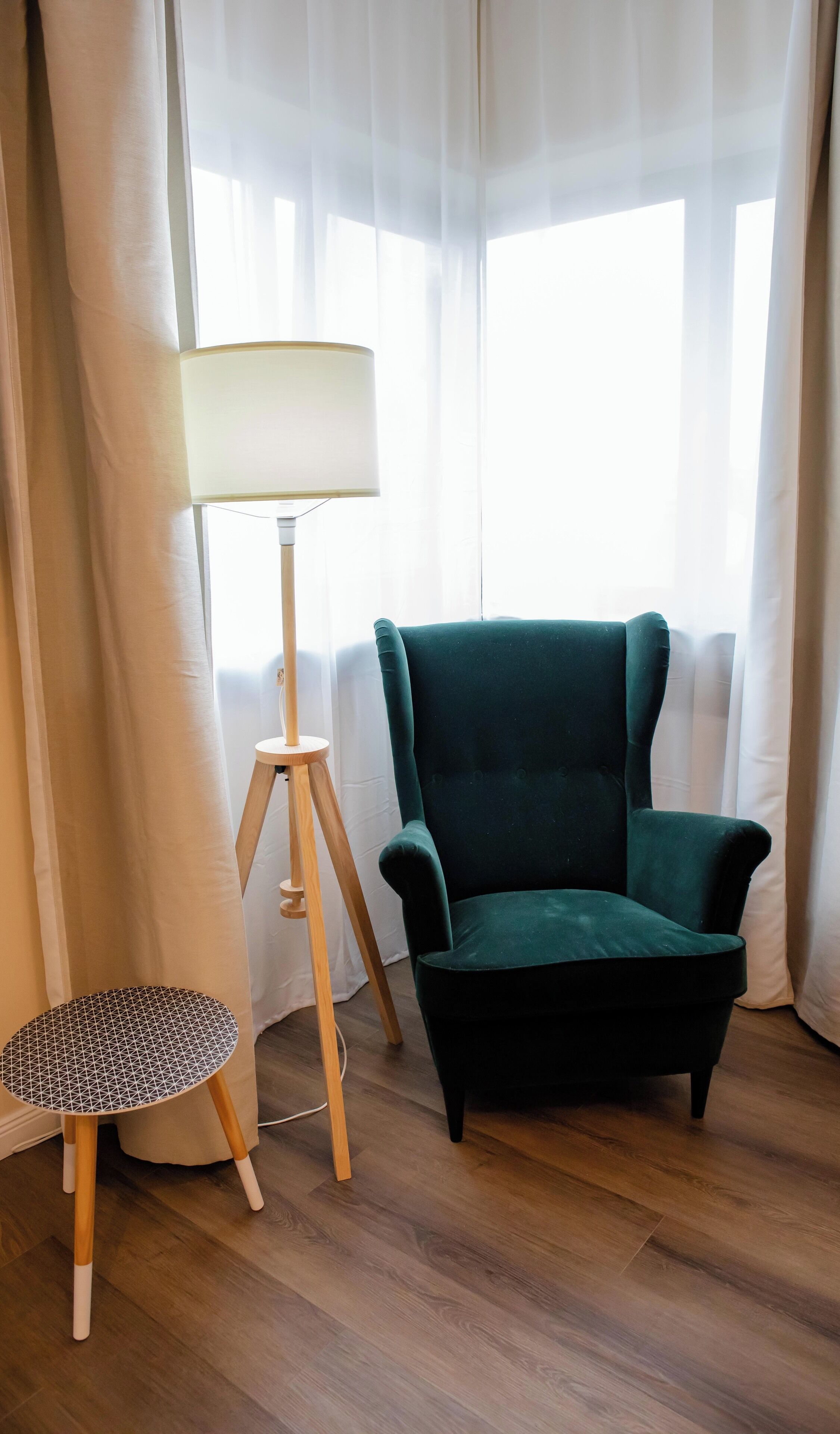 Um quarto com uma poltrona verde escuro, uma mesa redonda pequena com padrão geométrico e um abajur com cúpula branca.