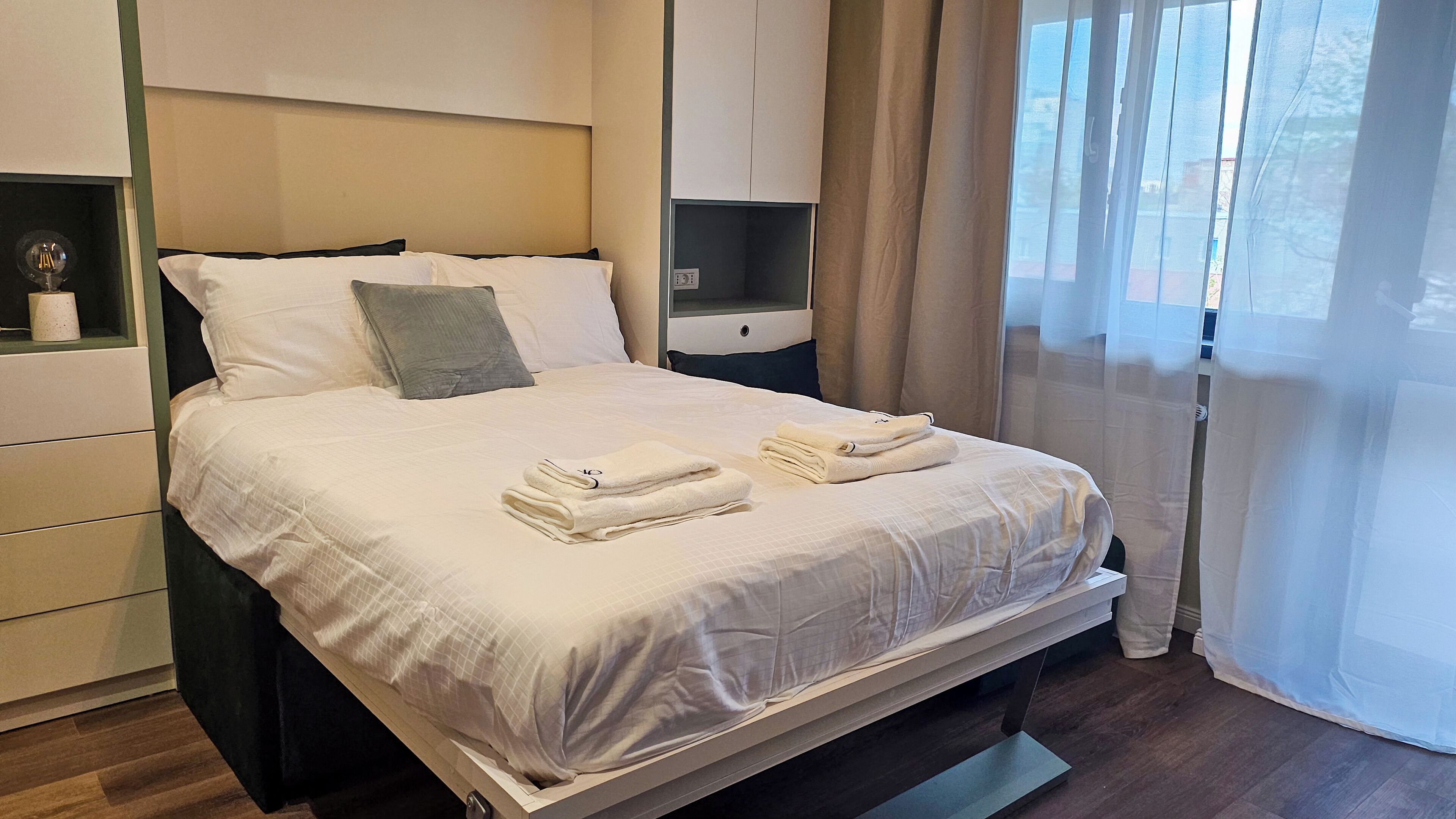 A neatly made bed with white linens and two folded towels, a bedside table with a lamp, and a window with sheer curtains.