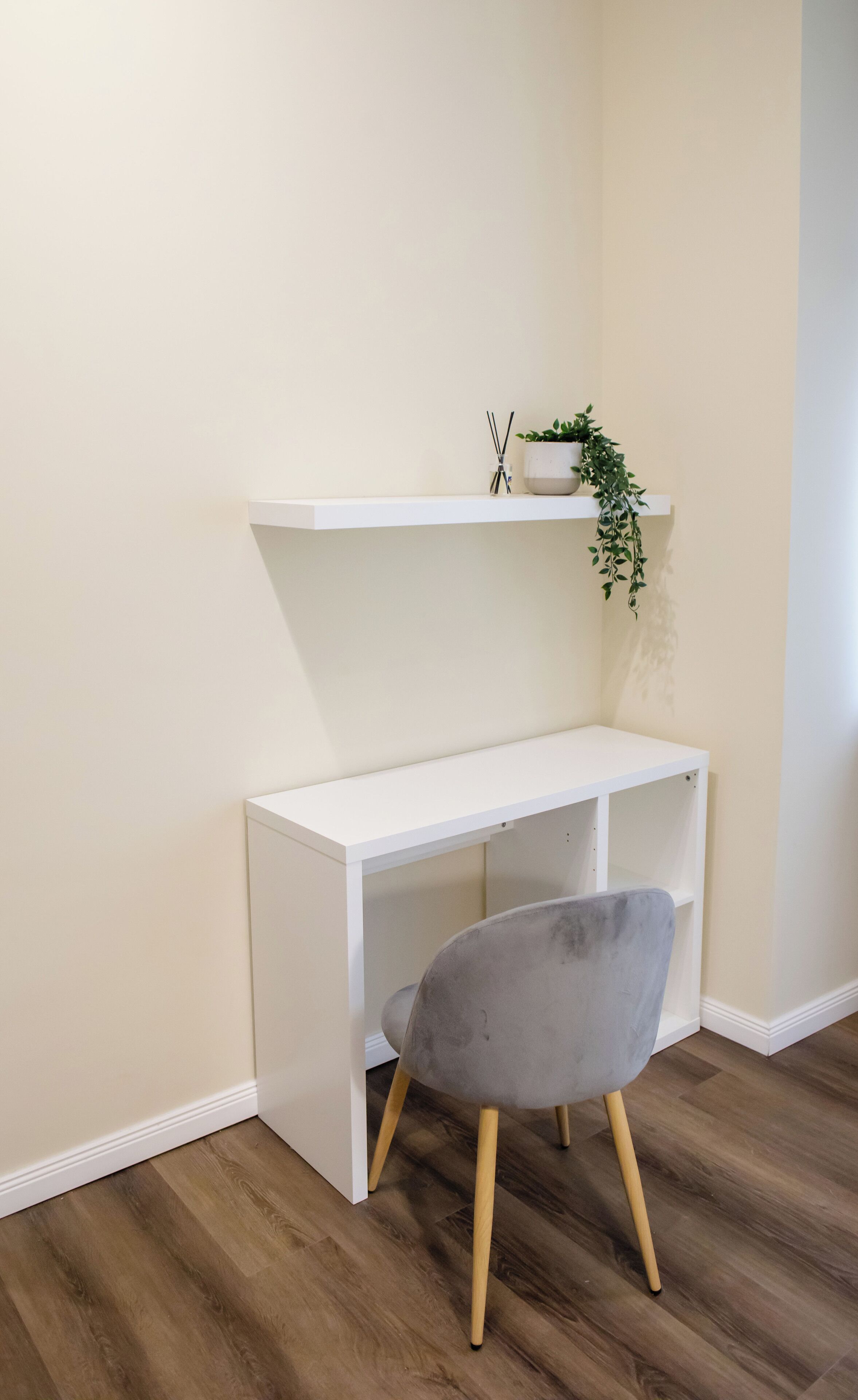 Una habitación de esquina con un escritorio blanco, una silla gris y una estantería con una planta.