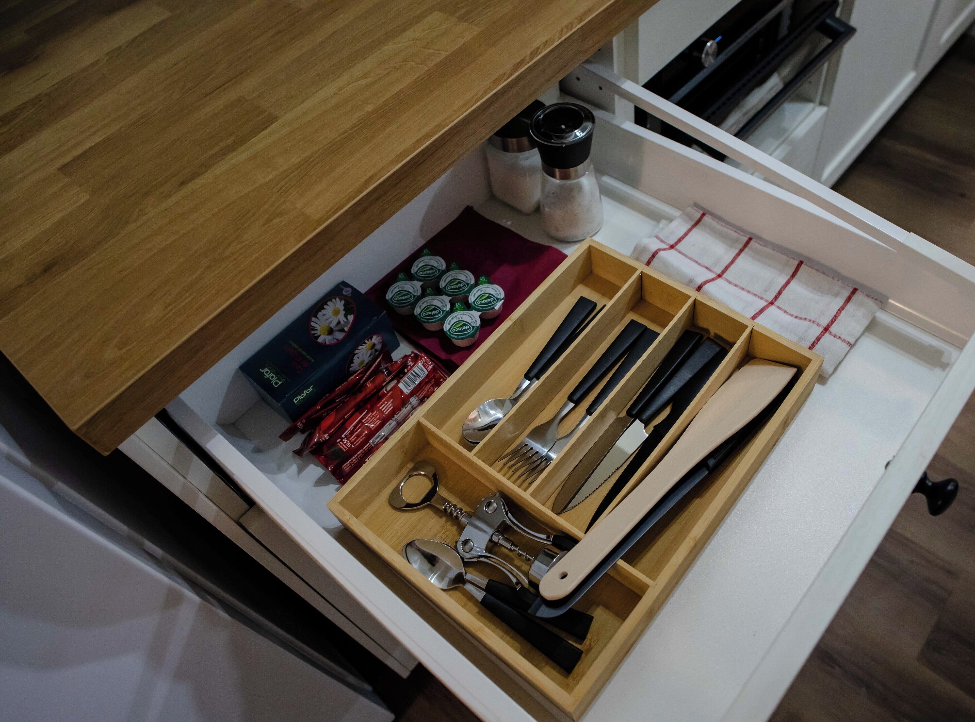 Un cajón de cocina con utensilios, una caja de cápsulas de café, saleros y pimenteros, y una tabla de cortar.