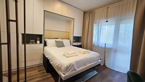 A modern hotel room with a large bed, a nightstand, a wall-mounted shelf, and a window with sheer curtains. - Little Bucharest downtown apartments (Bucharest)