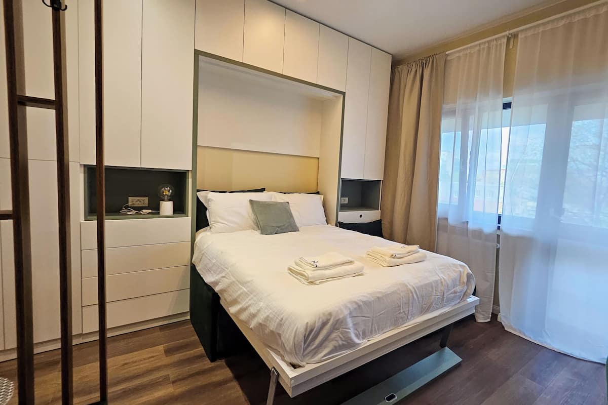 A modern hotel room with a large bed, a nightstand, a wall-mounted shelf, and a window with sheer curtains.