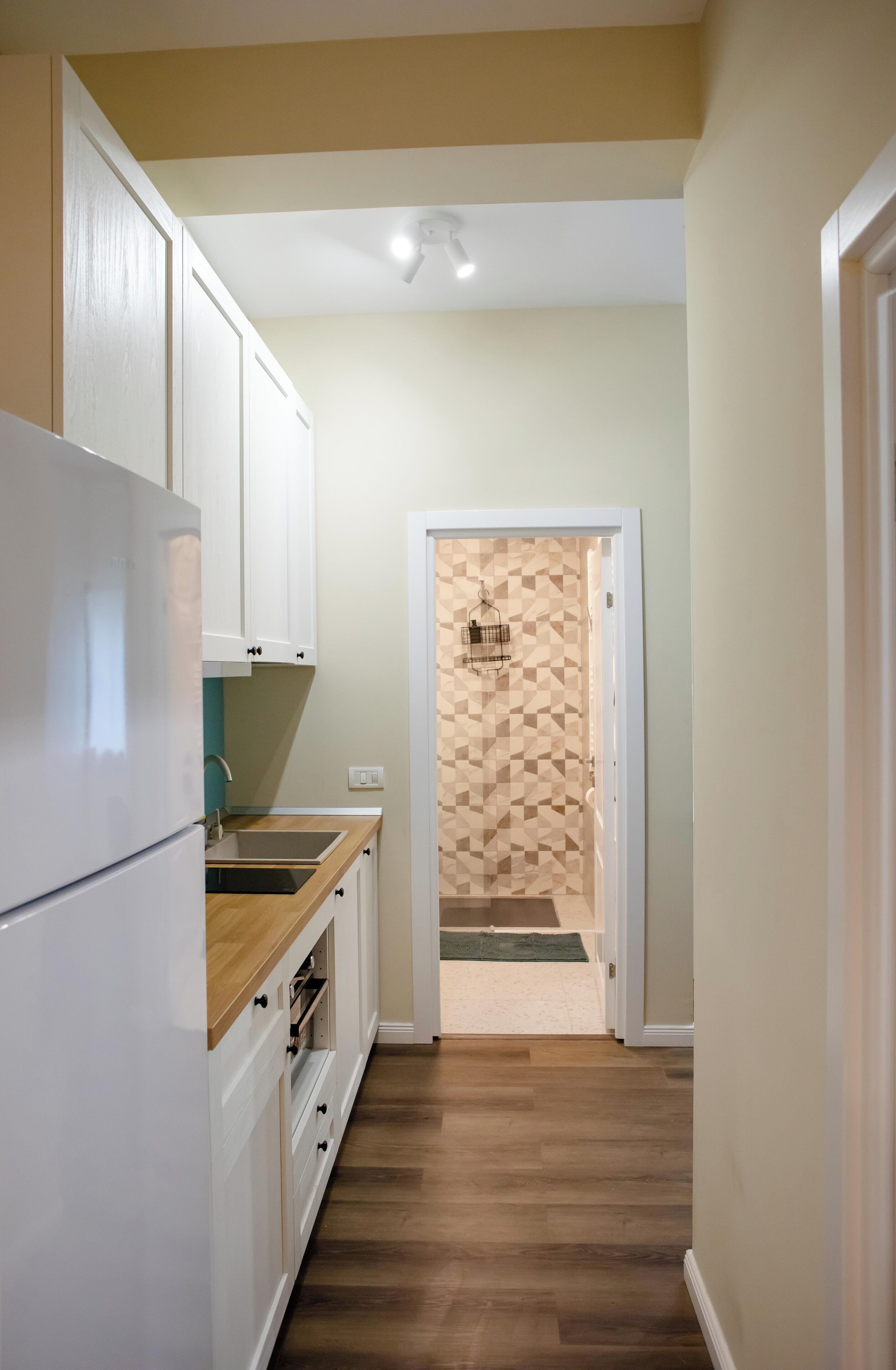 A narrow kitchen with white cabinets, a wooden countertop, and a built-in oven. A door leads to a room with a geometric-patterned wall.