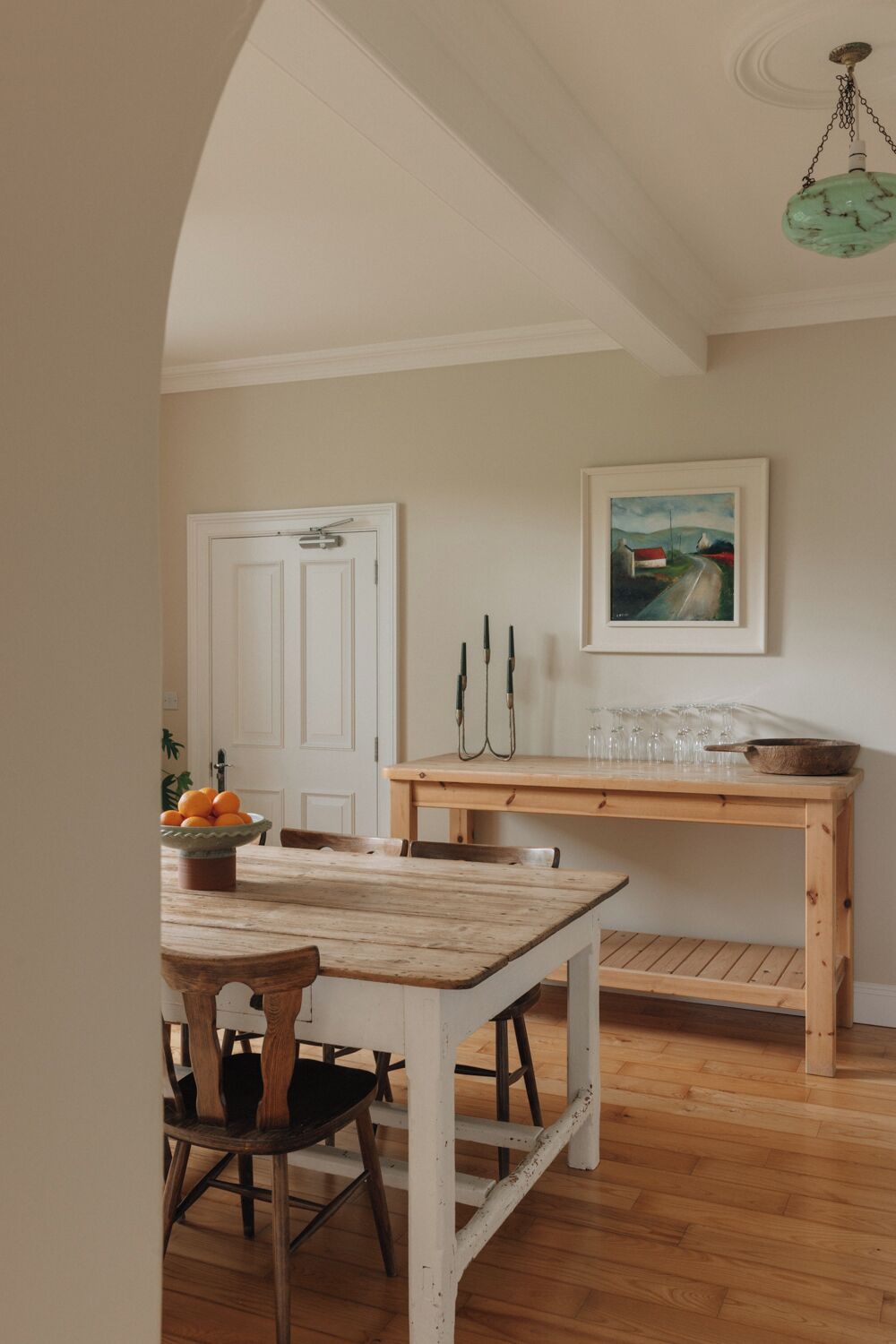 A rustic dining area with a wooden table and chairs, a white door, and a painting on the wall.