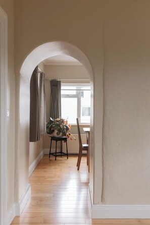 Een gang met een houten vloer, een gewelfde deuropening, een tafel met een plant en een stoel.