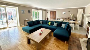 Un salon spacieux avec un grand canapé d’angle, une table basse en bois et un coin repas comprenant une table et des chaises blanches.