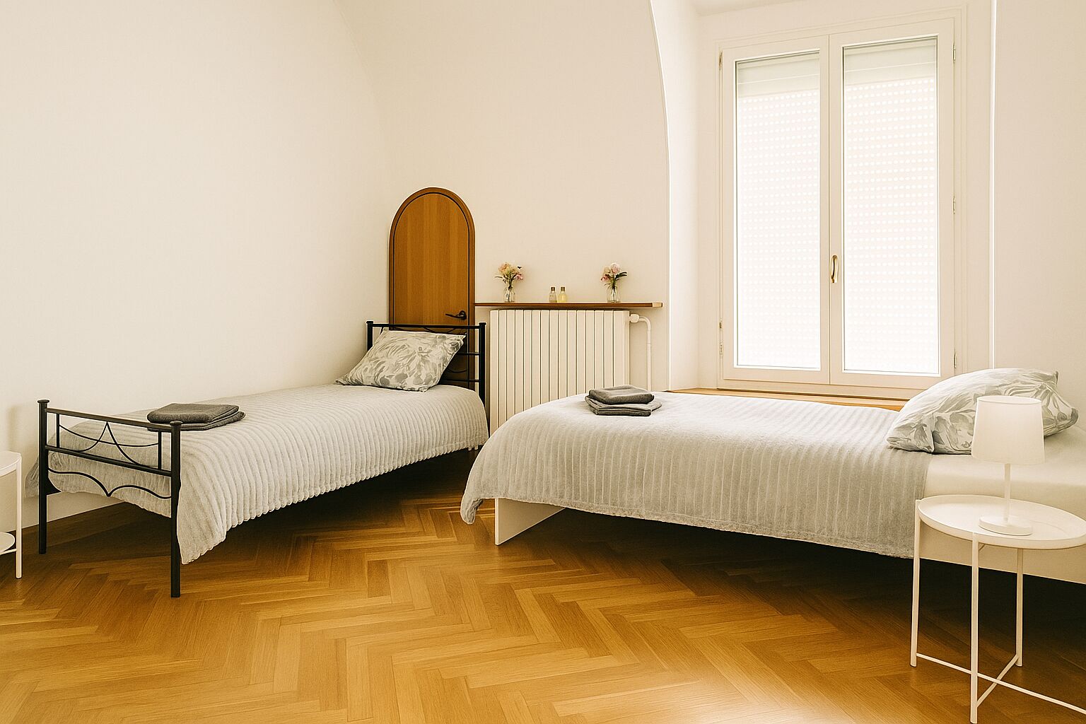 A bedroom with two single beds, a radiator, a window with blinds, a bedside table with a lamp, and a wooden floor.
