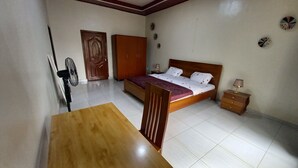 A bedroom with a wooden bed, a nightstand, a table, a fan, and a wardrobe. - PEACE & QUIET CORNER (Kigali)