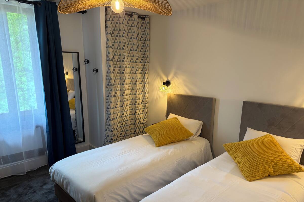 A hotel room with two beds, a large window with curtains, a patterned wall, and a ceiling light fixture.