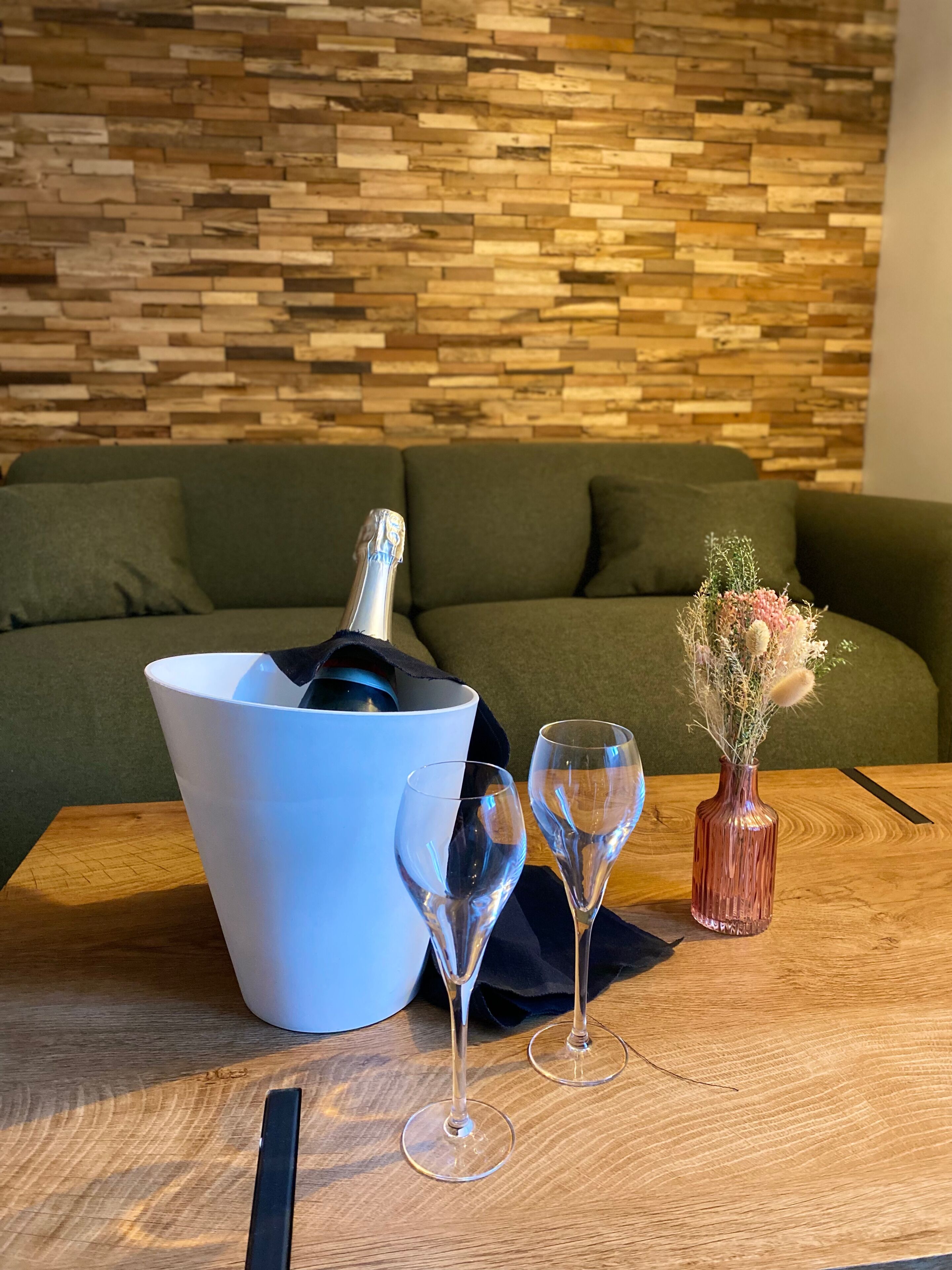 Una mesa de madera con un cubo blanco, dos copas de vino, un jarrón con flores secas y una botella de champán.