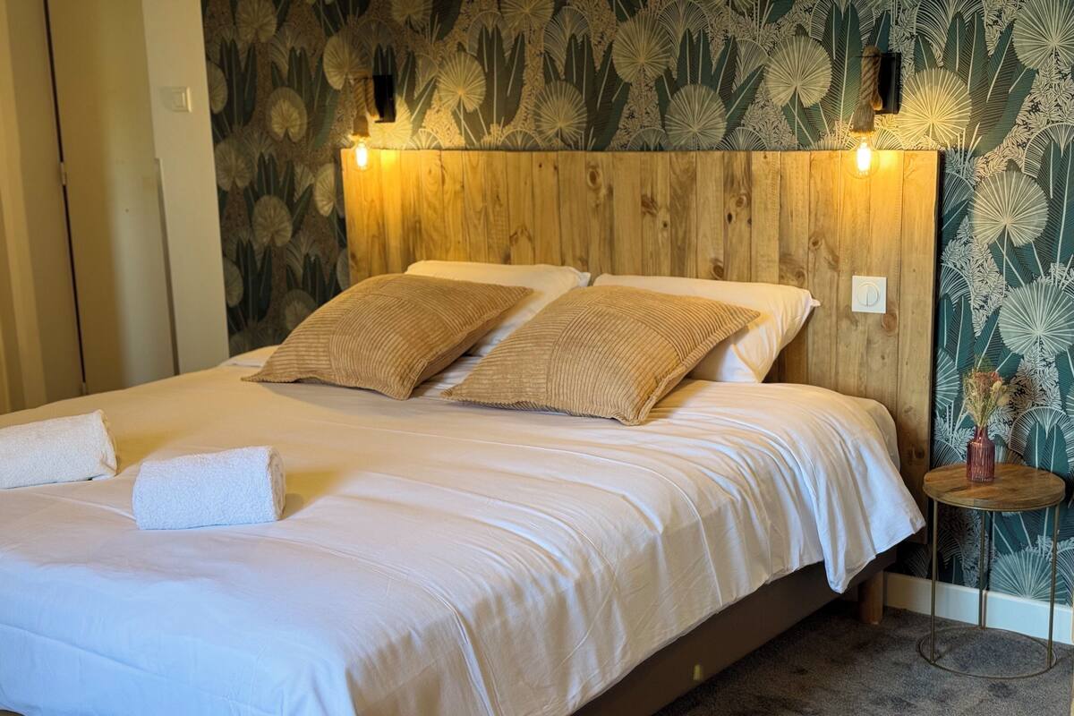 A bedroom with a wooden headboard, two beige pillows, a white bedspread, and a small round table with a vase.