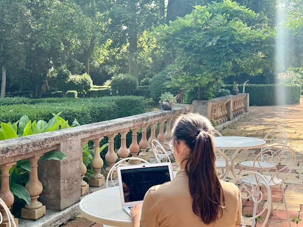 Una persona usando una computadora portátil en una mesa en un jardín con balaustrada de piedra y follaje.