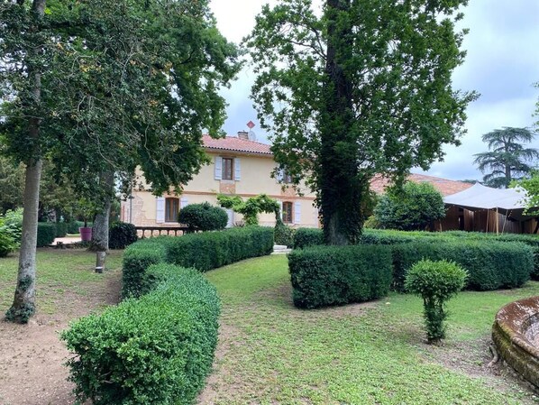 Un jardín bien cuidado con setos recortados, un árbol grande y un edificio con techo de tejas rojas.