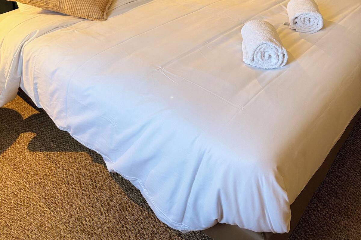 A neatly made bed with white linens and two rolled towels, a dark headboard, and a wall-mounted air conditioner.