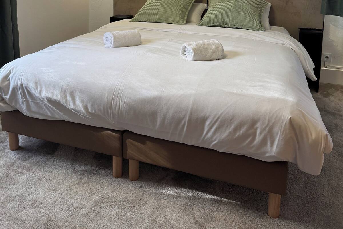 A neatly made bed with white linens and two rolled towels, a wooden headboard, and a small wall-mounted light fixture.