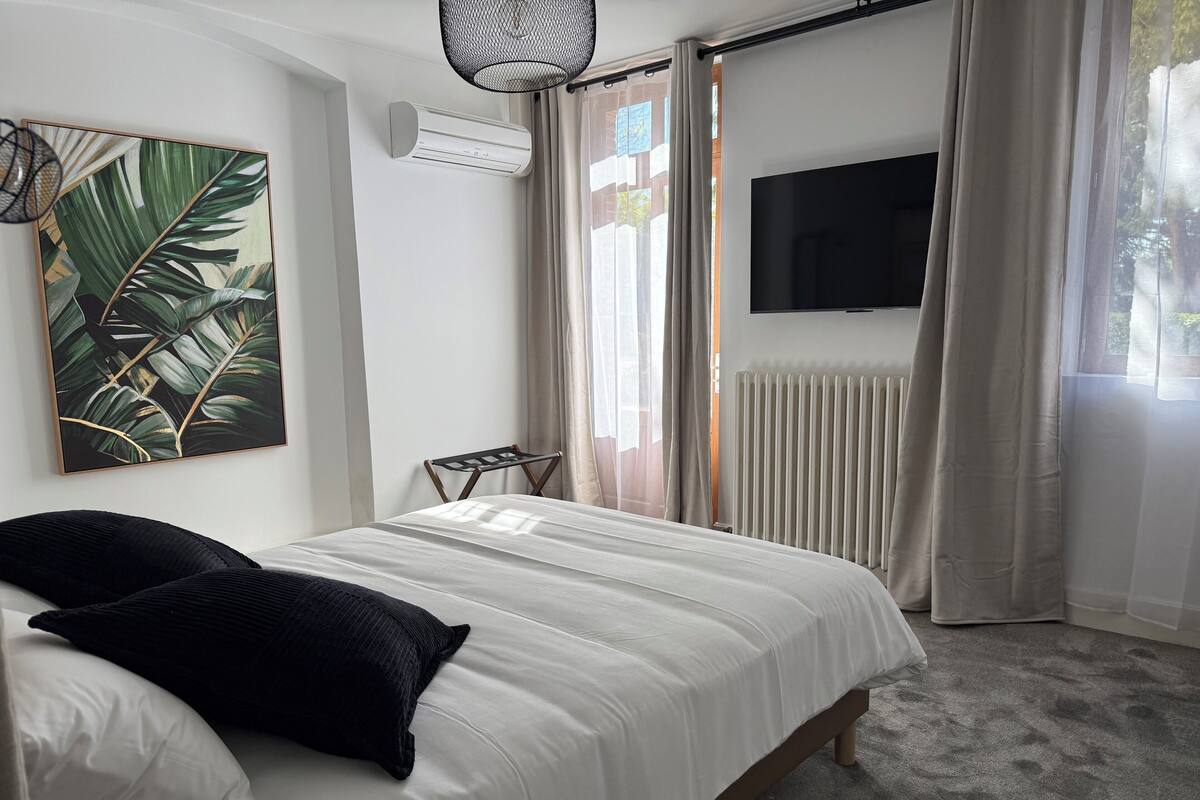A bedroom with a bed, a mounted TV, an air conditioning unit, a radiator, and a window with curtains.