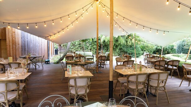 An outdoor dining area with tables and chairs under a canopy, surrounded by wooden walls and greenery. - hotel restaurant LEs Boissières (Bioule)