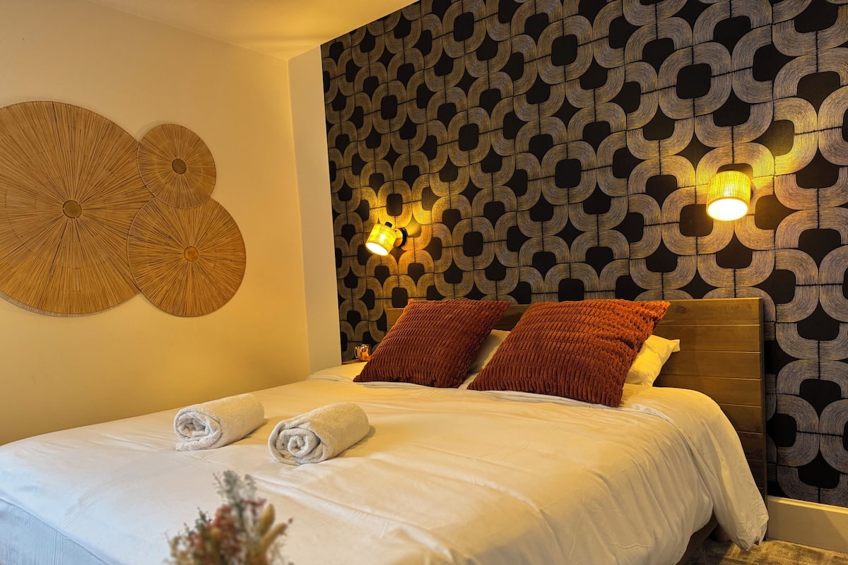 A neatly made bed with two rolled towels, a wall with a geometric pattern, and two wall-mounted lamps.