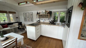 A modern kitchen with white cabinets, a wooden countertop, and a built-in stove. There is a dining area with a white table and chairs, and a window with a view of greenery outside. - Tiny house with terrace, beach chair and garden near Zingst (Pruchten)