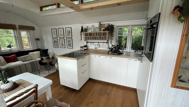 A modern kitchen with white cabinets, a wooden countertop, and a built-in stove. There is a dining area with a white table and chairs, and a window with a view of greenery outside. - Tiny house with terrace, beach chair and garden near Zingst (Pruchten)