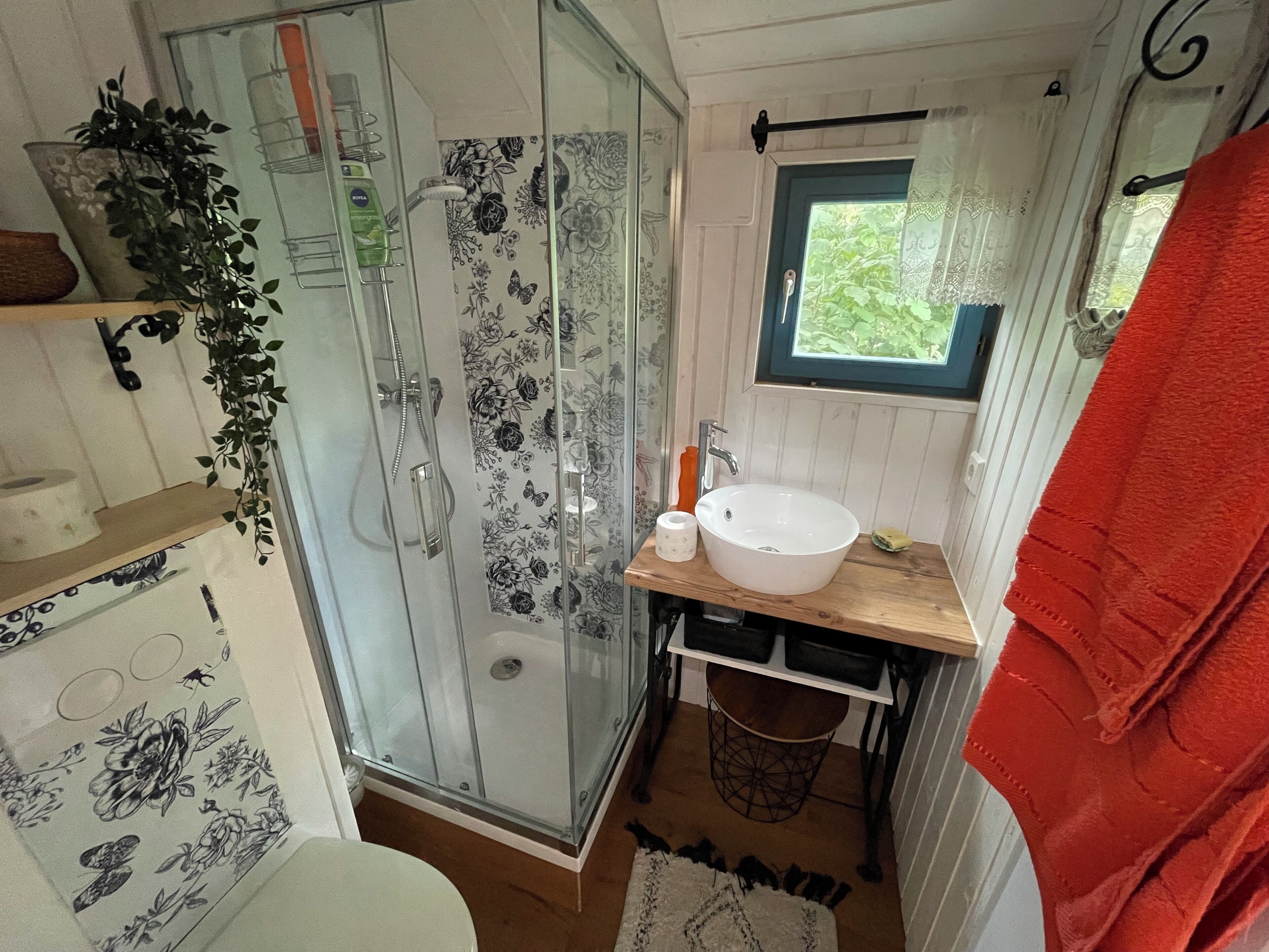 Een badkamer met een douchecabine van glas, een witte wastafel en een raam met uitzicht op groen.