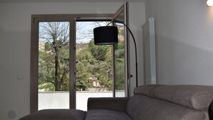 Ein Wohnzimmer mit einer Couch und einem Glastisch, einer schwarzen Stehlampe und Blick auf Bäume und Gebäude durch eine Glastür.