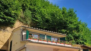 Ein zweigeschossiges Gebäude mit Balkon, davor ein rotes Auto und ein Hügel mit Bäumen.