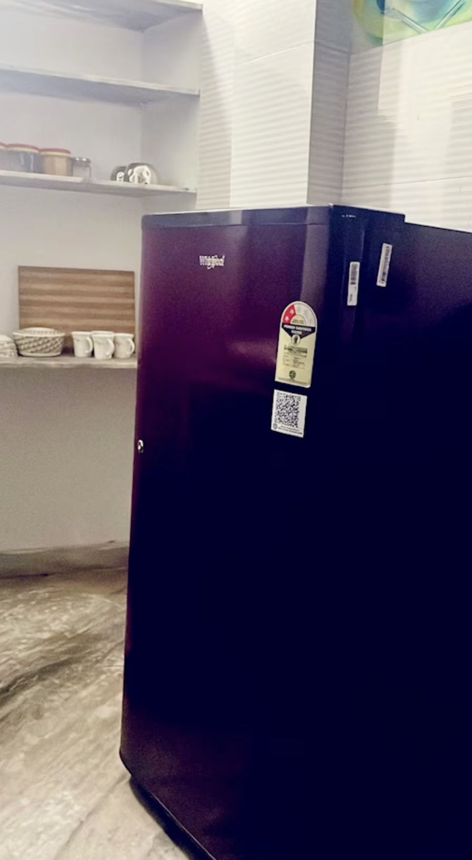 Un refrigerador de color oscuro con una etiqueta, ubicado en una habitación con una pared blanca y un estante con varios objetos.