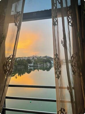 Vista a través de una ventana con cortinas ornamentales, reflejando un paisaje urbano y una masa de agua al atardecer.